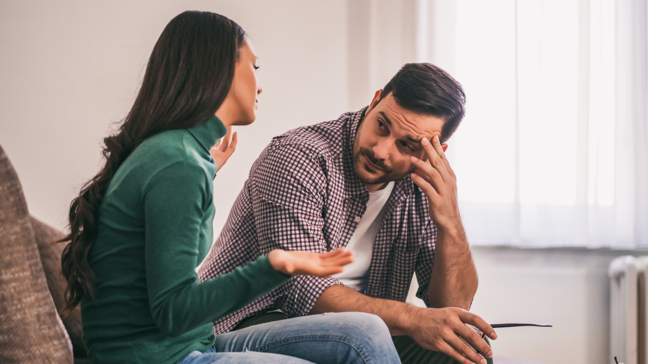 Man with a checked shirt rubs his forehead while listening to a woman speak in a bright room, conveying stress or worry during a discussion.