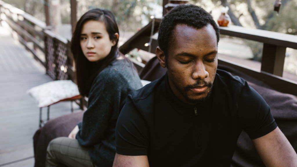 Man in a black shirt sits in foreground with eyes down, woman in gray sweater behind him looking away on a wooden deck outdoors.