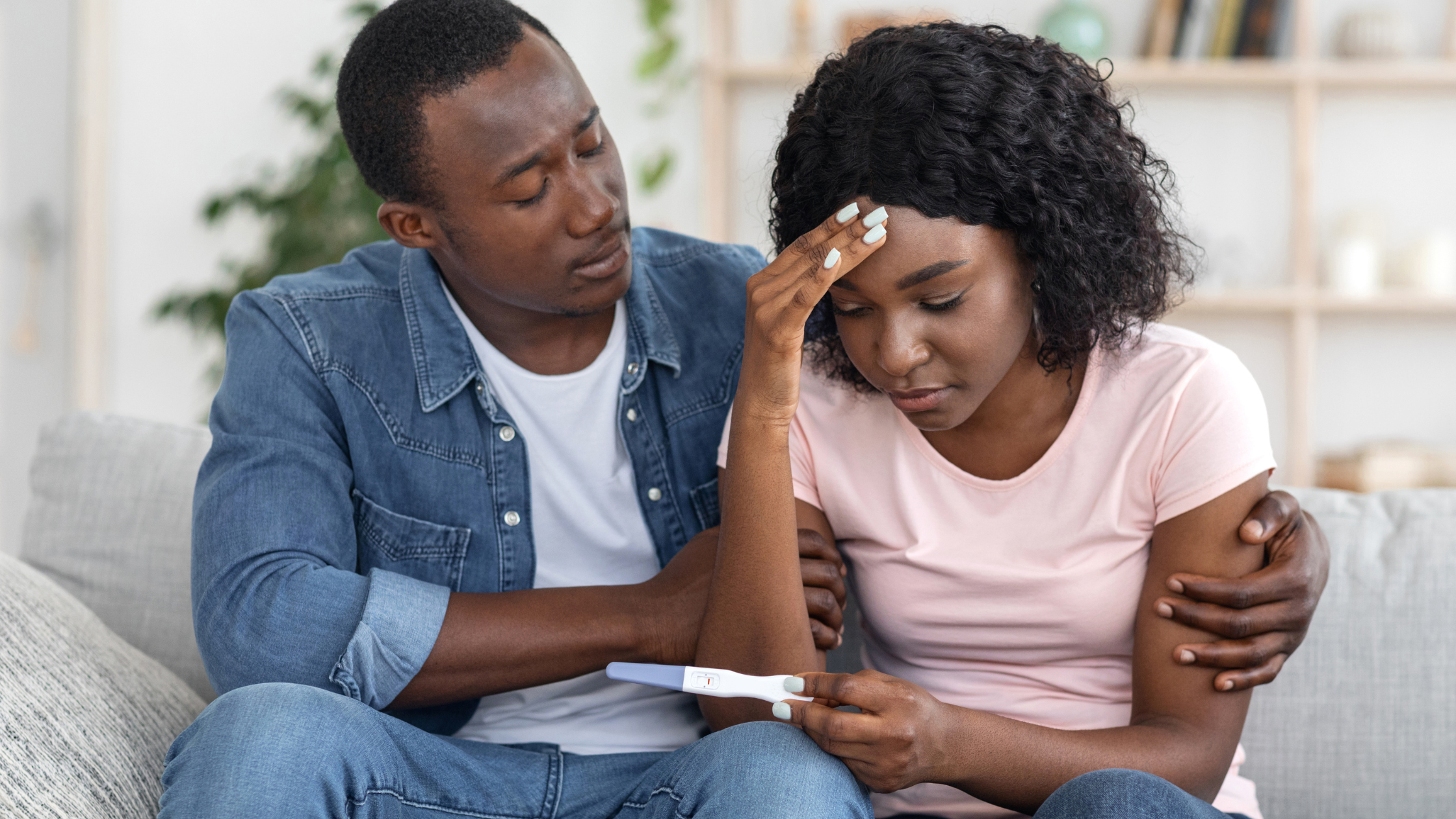 A couple sits on a sofa; the man comforts the woman who is holding a pregnancy test and looks concerned.